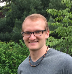 Photo of Andrew outside, with trees in the background. He wears glasses and a gray t-shirt.